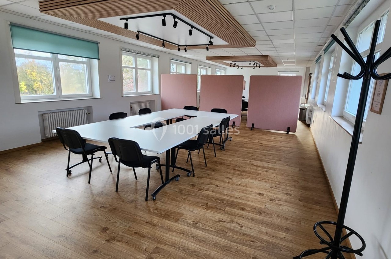 Salle de réunion lumineuse avec tables blanches, chaises noires, parquet en bois et cloisons roses en arrière-plan.