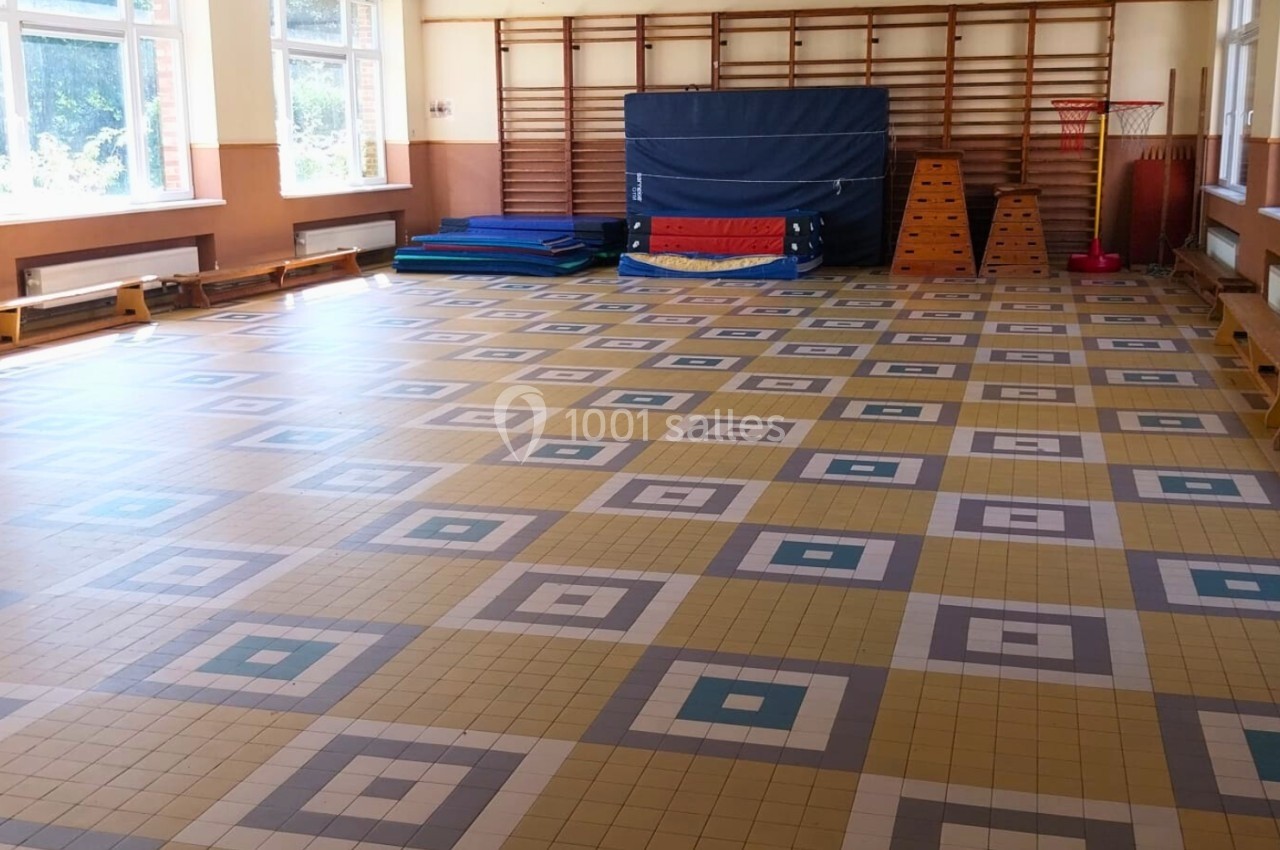 Salle de sport lumineuse avec sol en carrelage géométrique, tapis empilés et matériel d'exercice contre le mur.