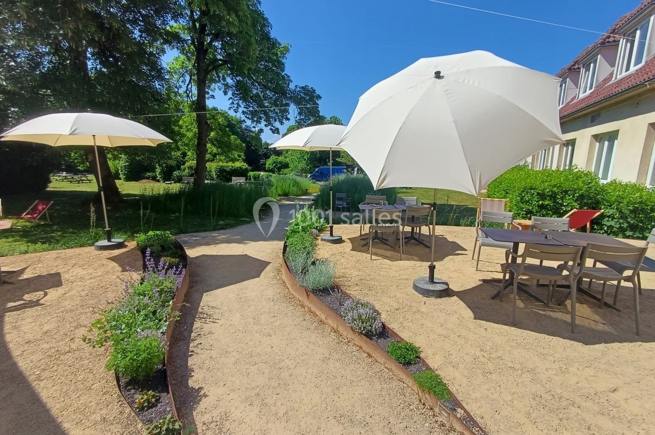 Terrasse extérieure avec tables, chaises et parasols, entourée de plantes et d'arbres sous un ciel ensoleillé.