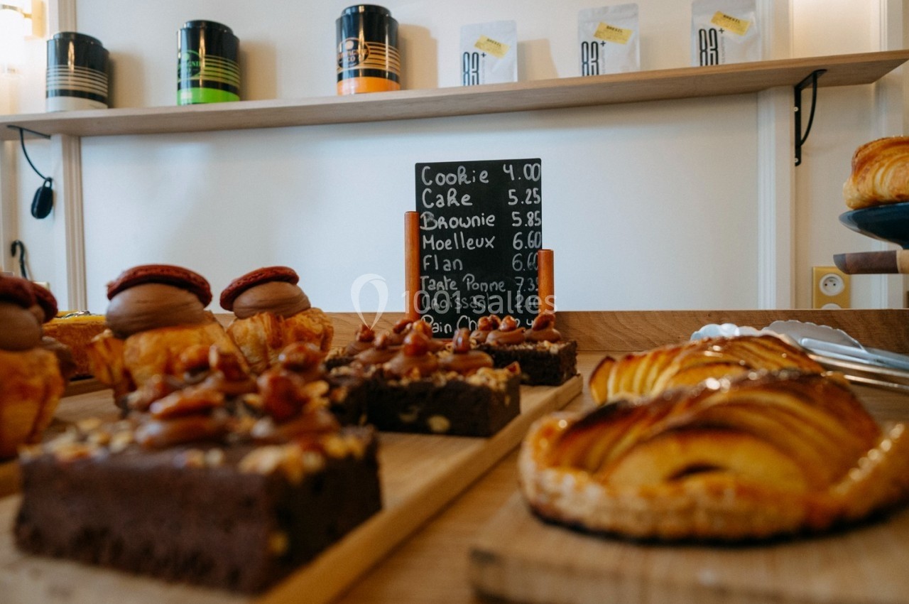 Pâtisseries variées sur un comptoir en bois avec une ardoise indiquant les prix en arrière-plan.