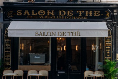 Façade d'un salon de thé parisien avec une devanture noire, une enseigne beige et des chaises en terrasse.