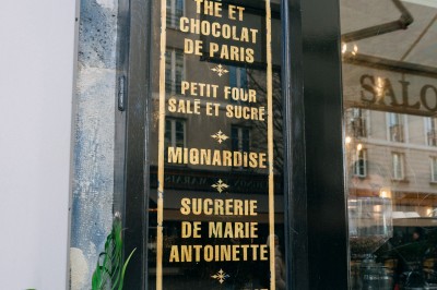 Façade d'un salon de thé parisien avec une devanture noire, une enseigne beige et des chaises en terrasse.
