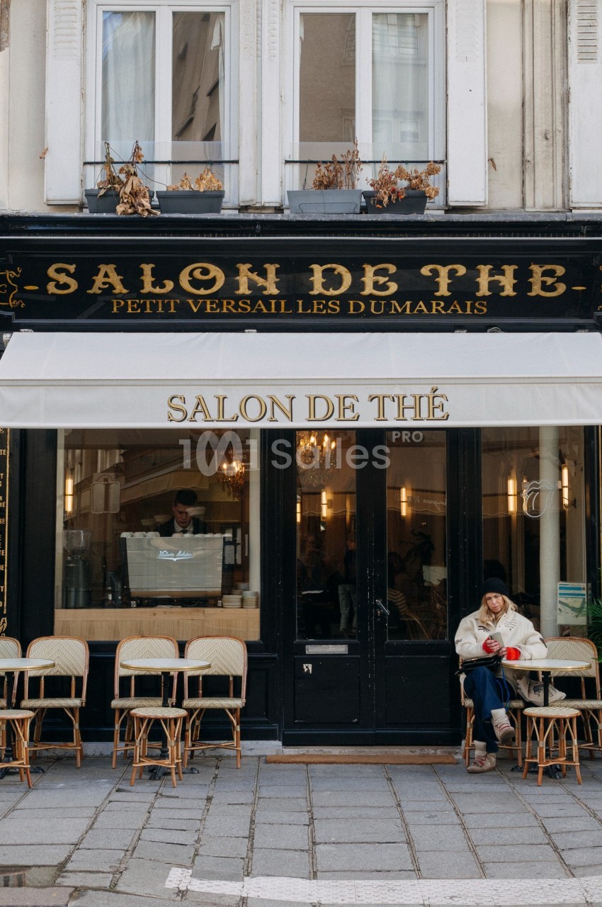 Façade d'un salon de thé parisien avec terrasse, chaises en osier et une personne assise à l'extérieur.