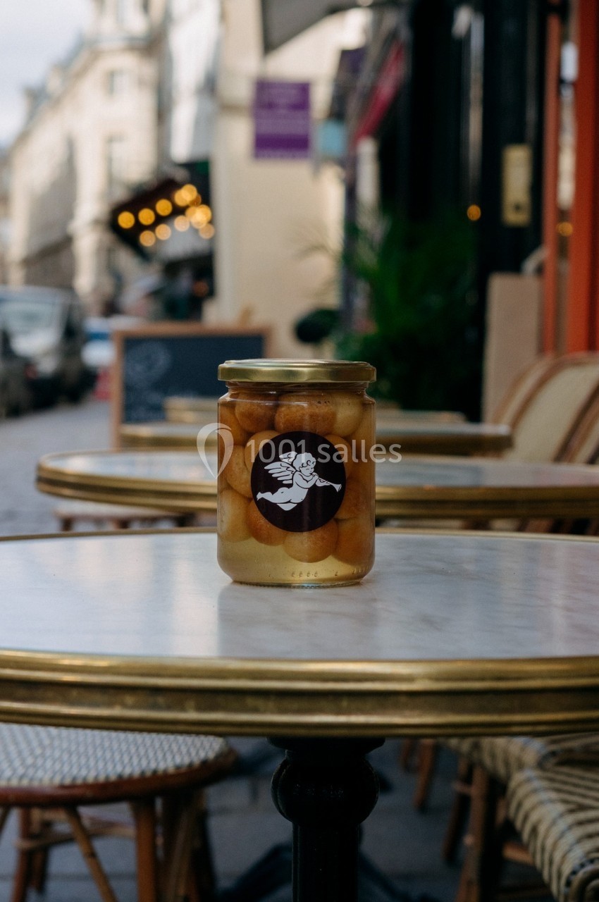 Bocal de légumes marinés posé sur une table en marbre dans une rue bordée de cafés et de boutiques.