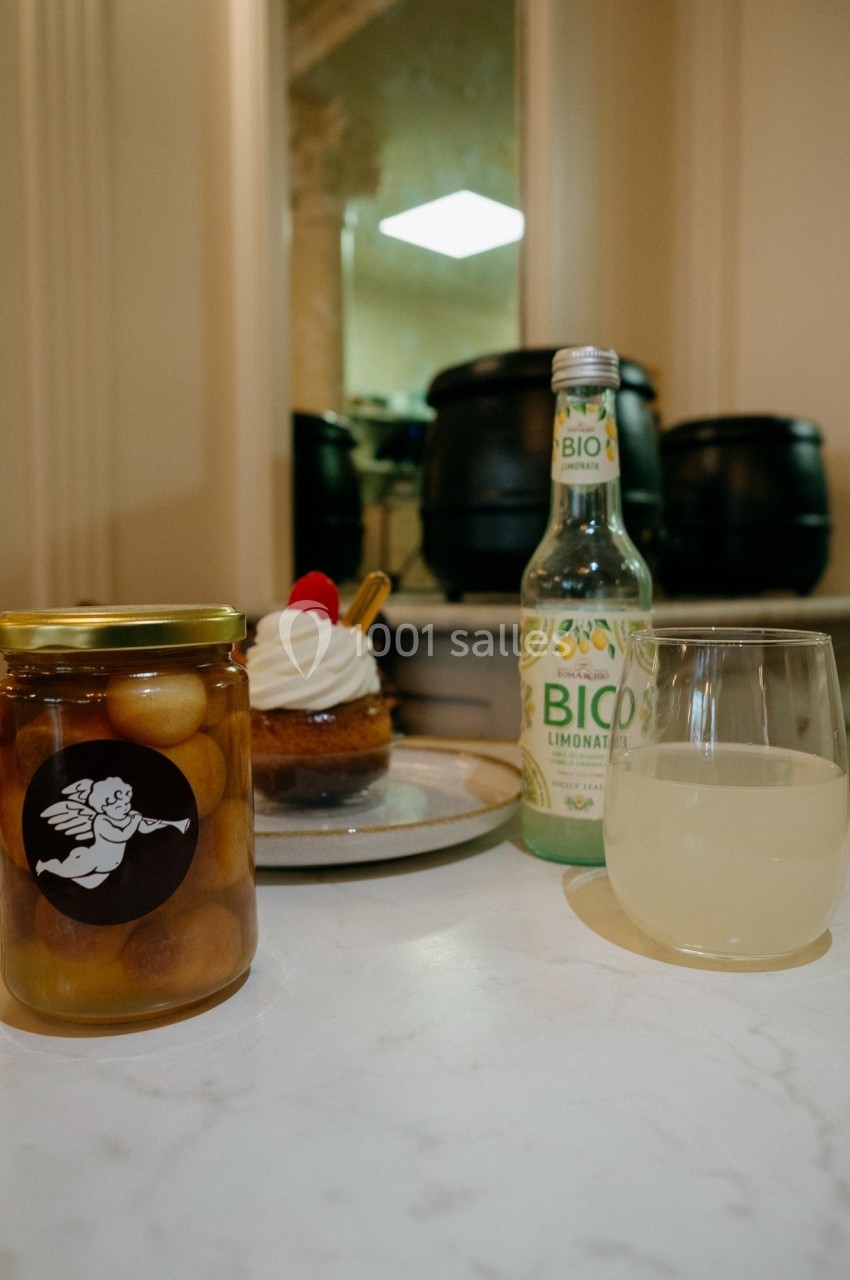 Un pot de fruits au sirop, une tartelette, une bouteille de limonade bio et un verre sur une table en intérieur.