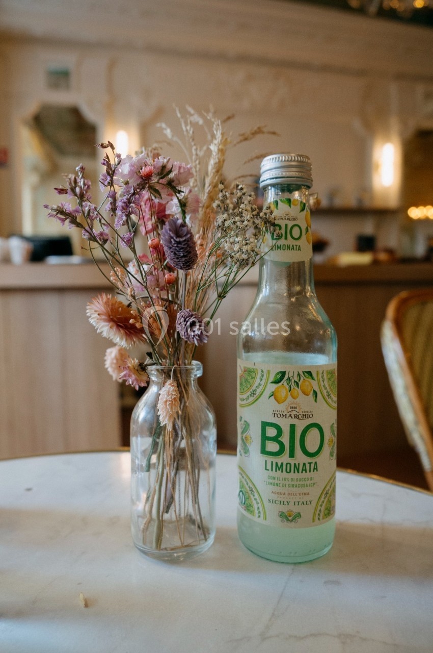 Bouteille de limonade bio posée sur une table en marbre à côté d'un vase contenant des fleurs séchées.