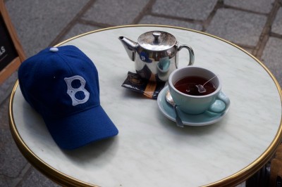 Façade d'un salon de thé parisien avec une devanture noire, une enseigne beige et des chaises en terrasse.