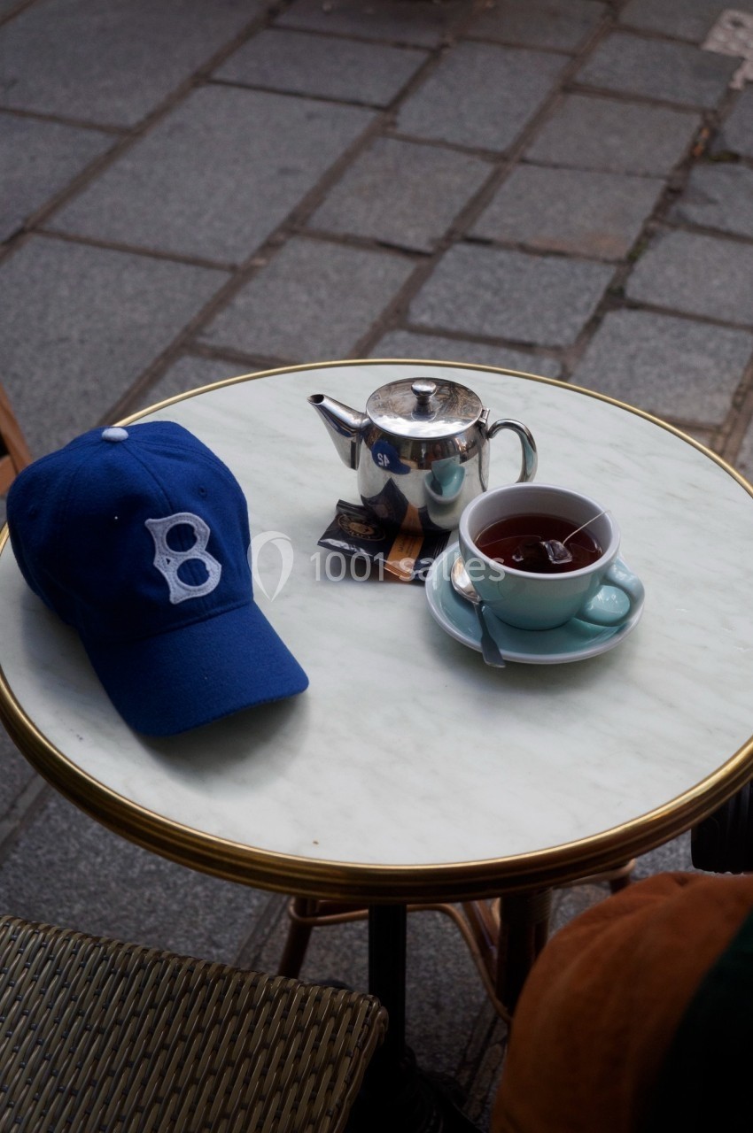 Casquette bleue, théière en métal et tasse de thé sur une table ronde en marbre, en extérieur sur un trottoir.