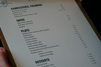 Façade d'un salon de thé parisien avec une devanture noire, une enseigne beige et des chaises en terrasse.