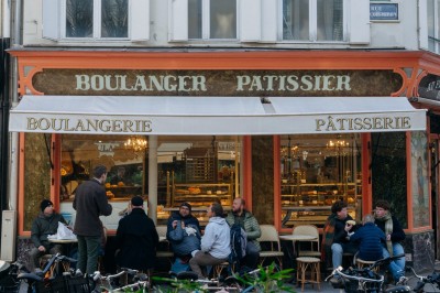 Façade d'un salon de thé parisien avec une devanture noire, une enseigne beige et des chaises en terrasse.