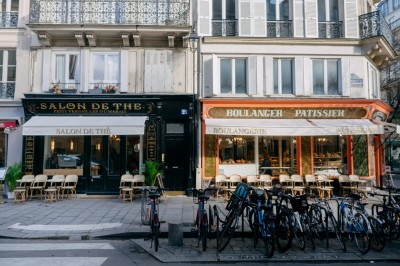 Façade d'un salon de thé parisien avec une devanture noire, une enseigne beige et des chaises en terrasse.