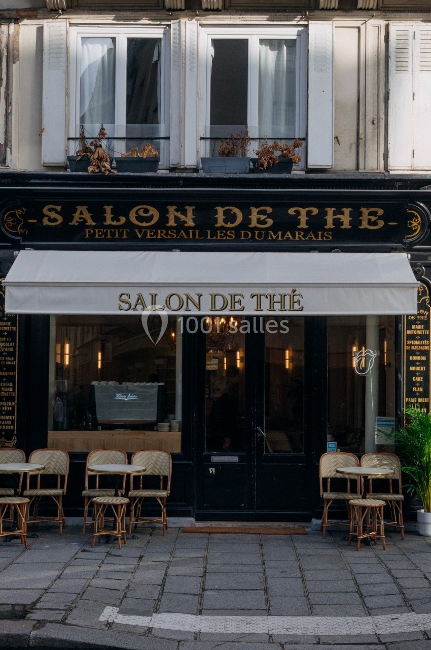 Façade d'un salon de thé parisien avec une devanture noire, une enseigne beige et des chaises en terrasse.