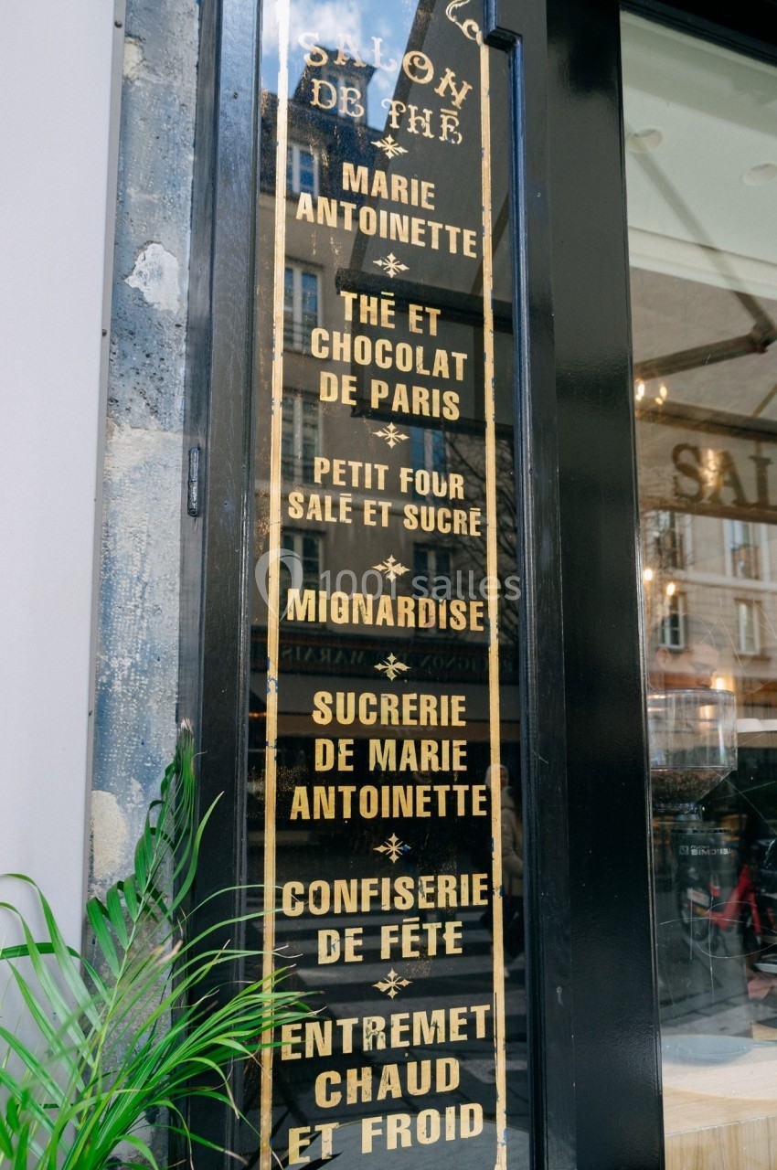 Enseigne verticale d'un salon de thé avec une liste de spécialités, reflet de bâtiments dans la vitre.