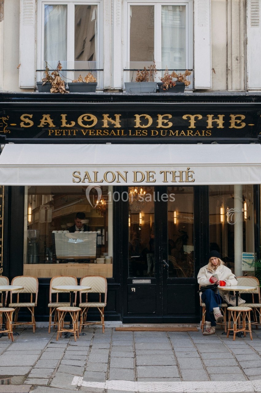 Façade d'un salon de thé parisien avec terrasse, chaises en osier et une personne assise à l'extérieur.
