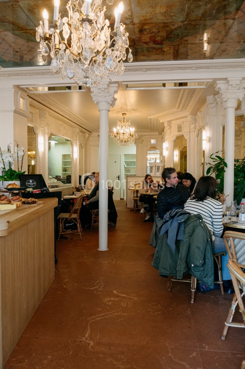 Intérieur d'un café lumineux avec des clients assis, un comptoir à pâtisseries et des lustres décoratifs.
