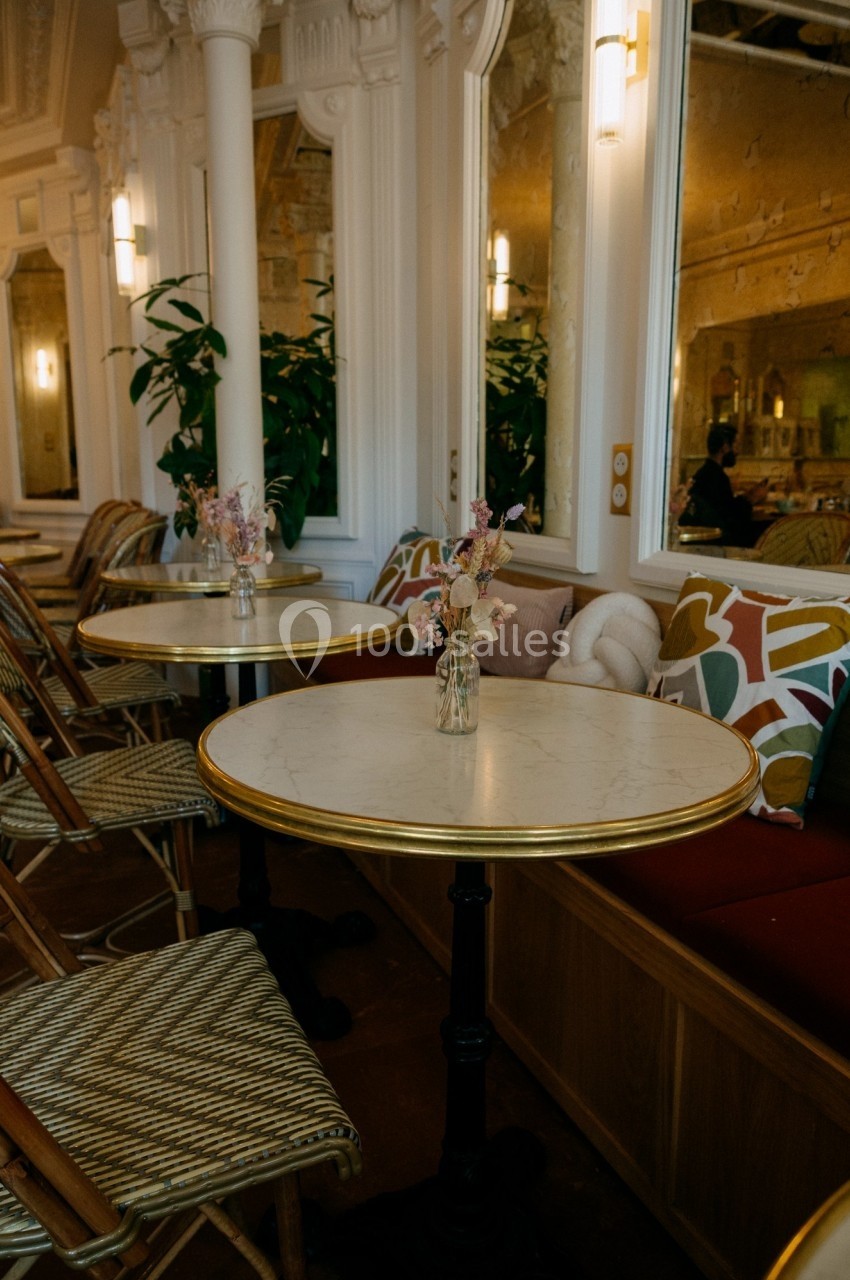Tables en marbre avec chaises en rotin dans un café élégant, décoré de coussins colorés et de fleurs en vase.