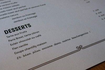 Façade d'un salon de thé parisien avec une devanture noire, une enseigne beige et des chaises en terrasse.