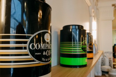 Façade d'un salon de thé parisien avec une devanture noire, une enseigne beige et des chaises en terrasse.