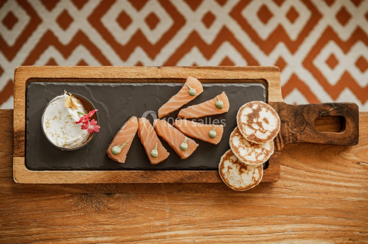Tranches de saumon fumé avec crème, blinis et décoration florale, présentés sur une planche en bois.