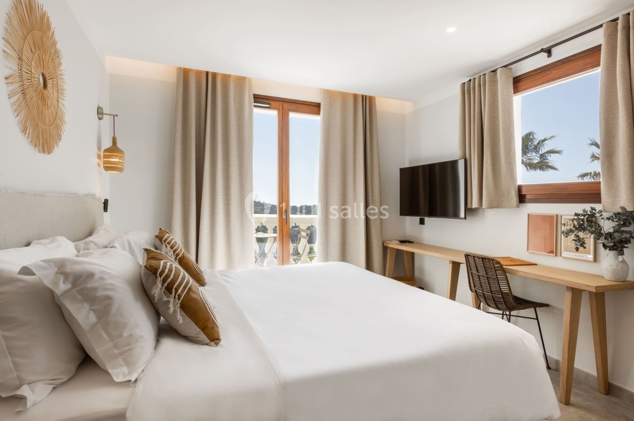 Chambre lumineuse avec lit double, bureau en bois, télévision murale et grandes fenêtres donnant sur un paysage extérieur.