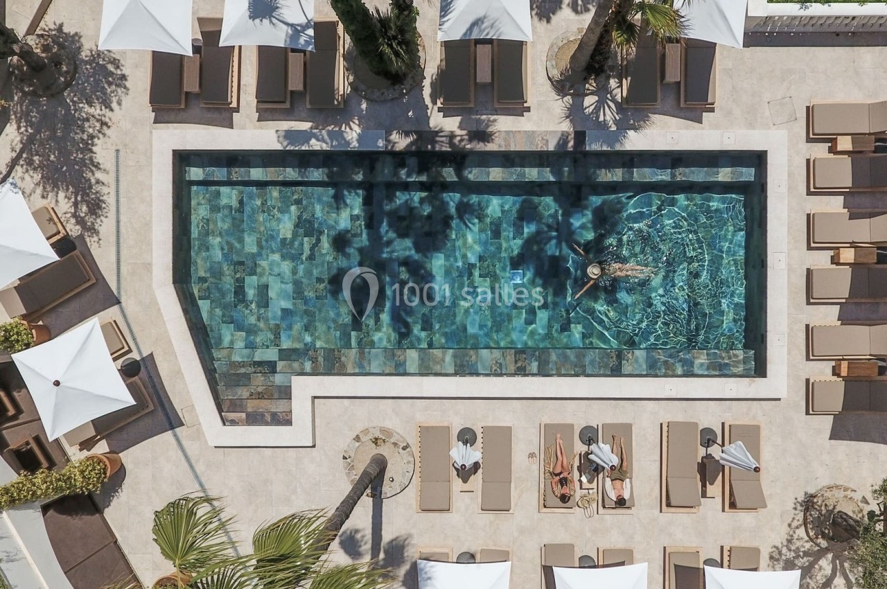 Vue aérienne d'une piscine entourée de transats, parasols blancs et palmiers, avec une personne nageant au centre.