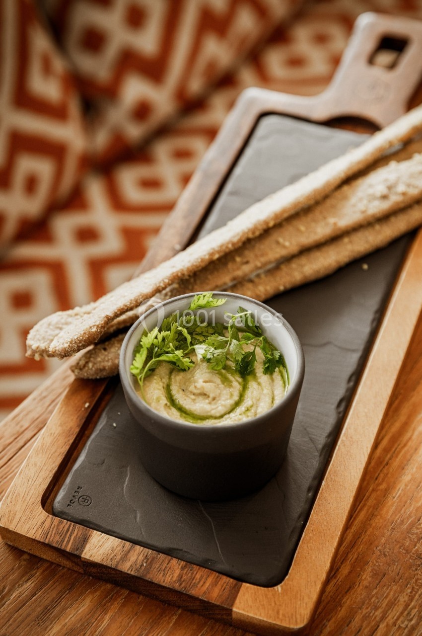 Bol de houmous garni d'herbes fraîches, accompagné de gressins sur une planche en bois.