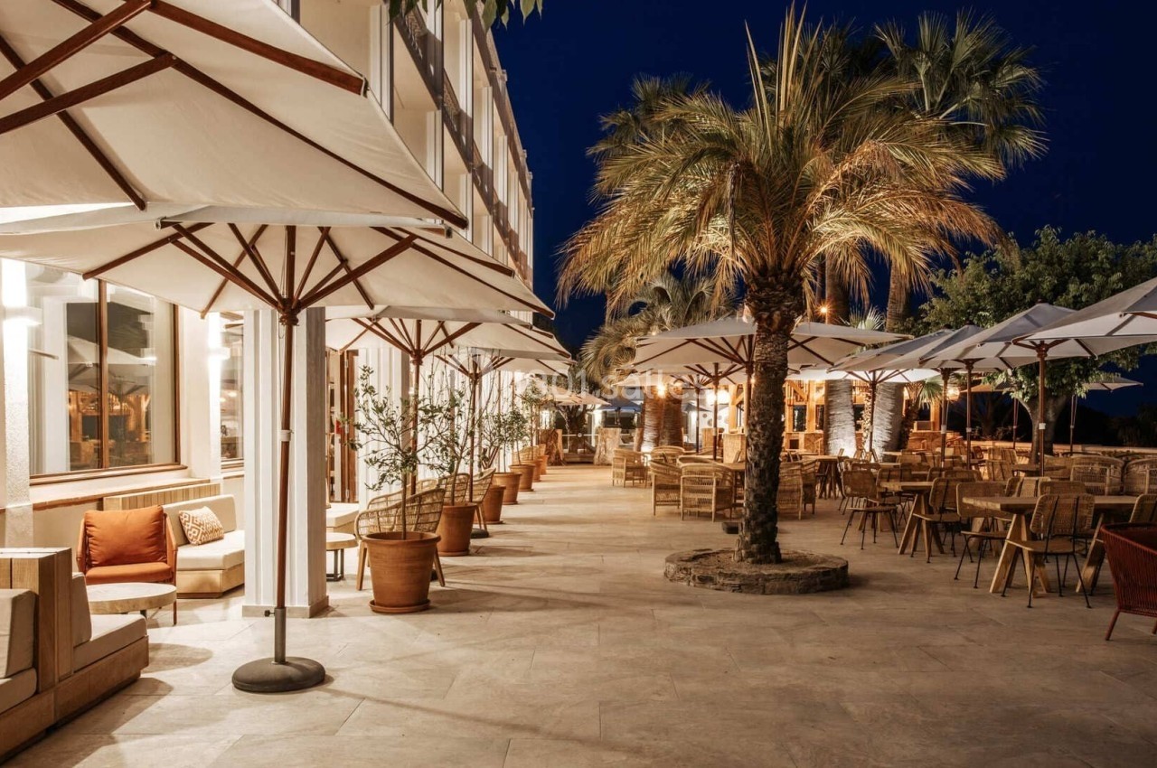 Terrasse d'un restaurant éclairée la nuit, avec des parasols, des tables, des chaises et des palmiers.