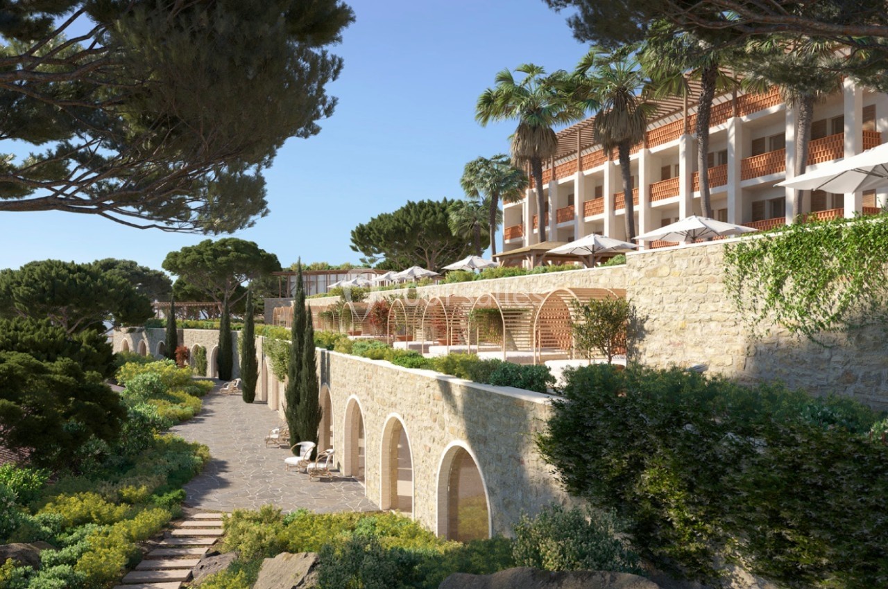 Chemin bordé de végétation menant à un bâtiment en pierre avec terrasses et parasols, entouré de pins et palmiers.