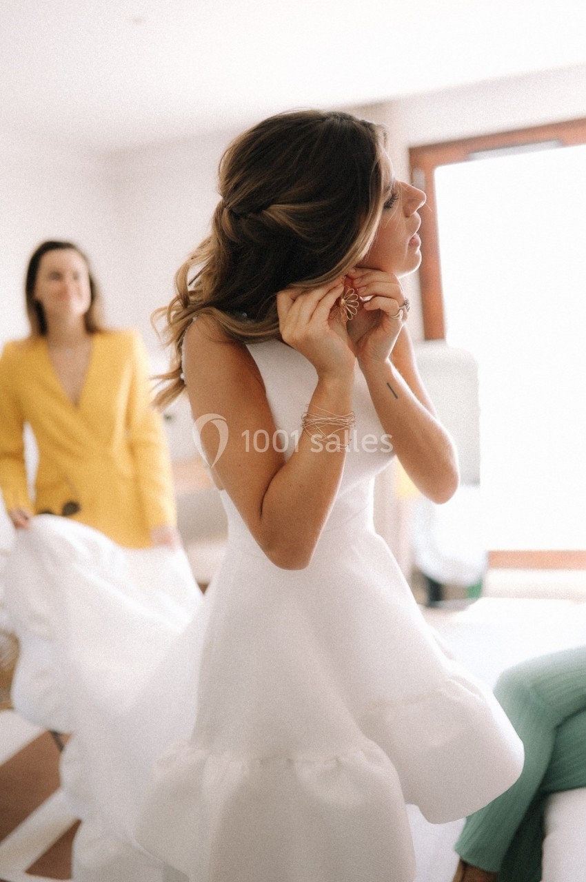 Une femme en robe blanche ajuste ses boucles d'oreilles, accompagnée d'une autre personne en veste jaune en arrière-plan.