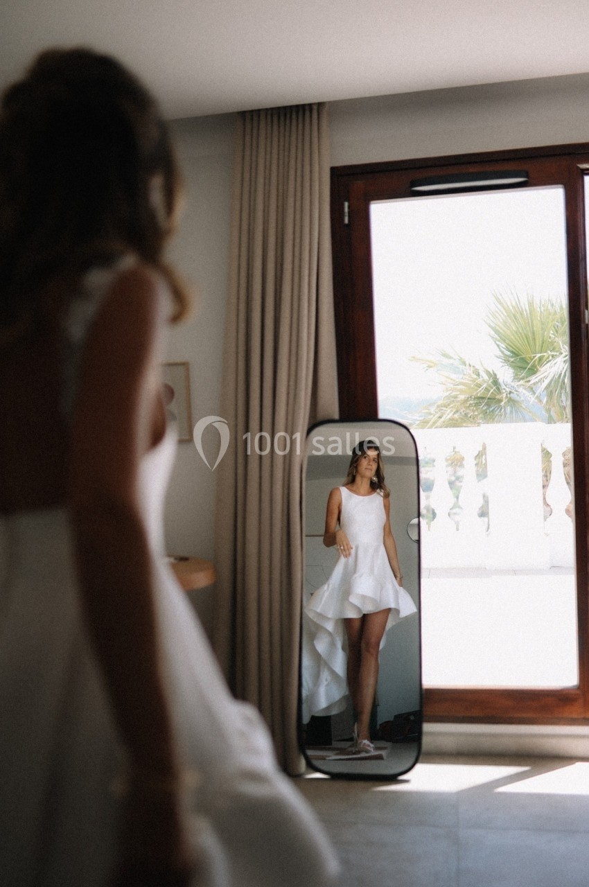 Une femme en robe blanche se regarde dans un miroir près d'une fenêtre laissant entrer la lumière naturelle.