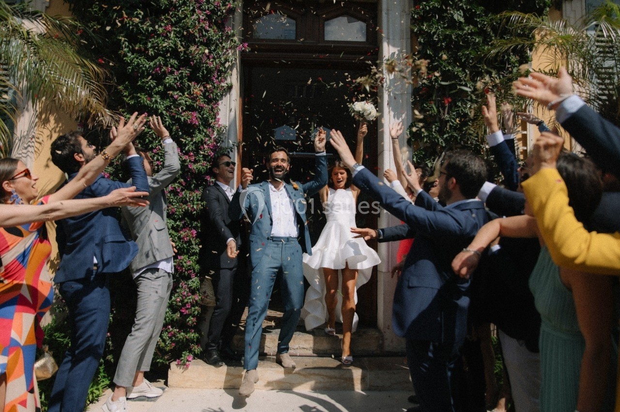 Un couple sort d'un bâtiment sous une pluie de confettis, entouré d'invités applaudissant joyeusement.