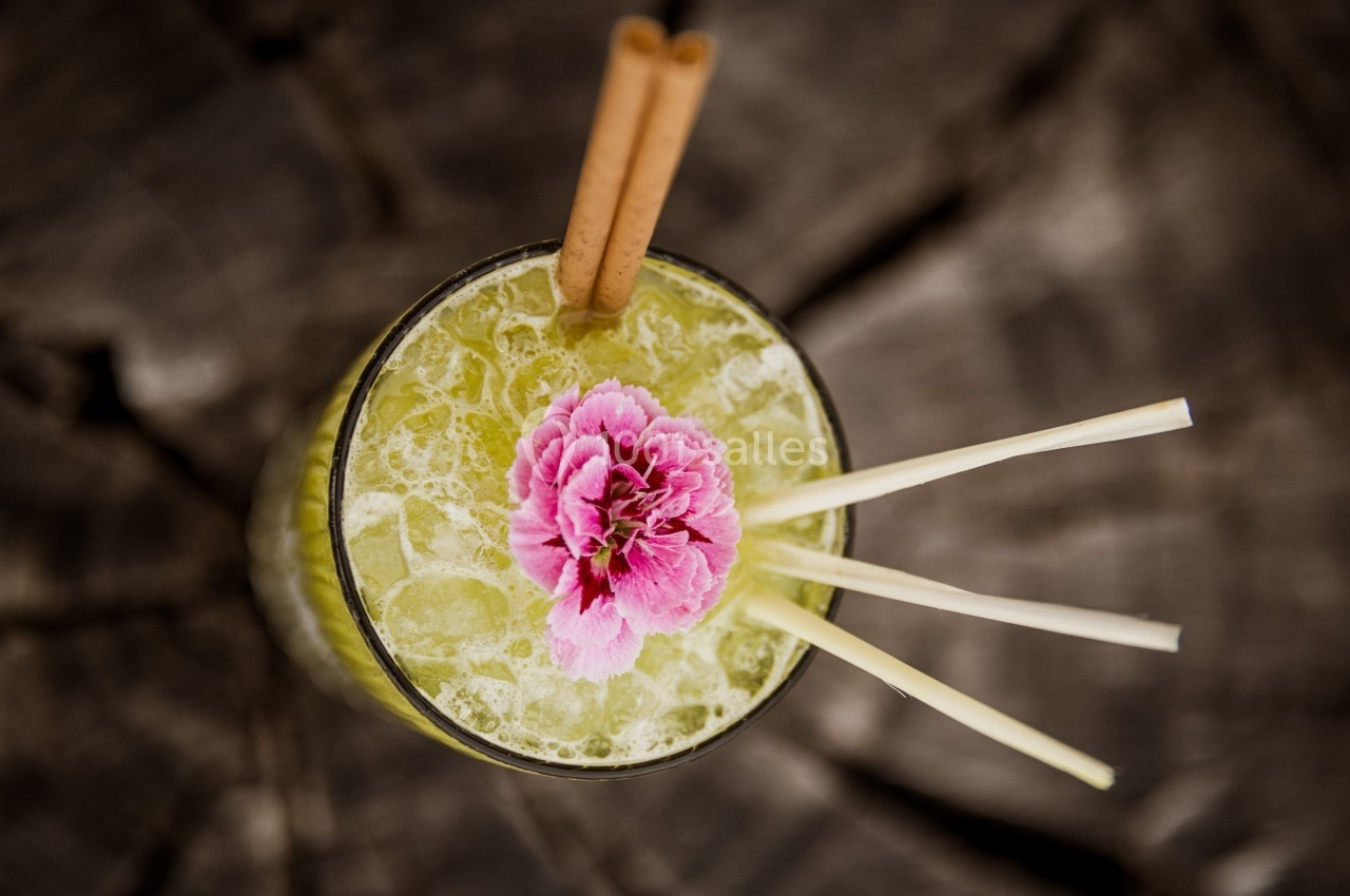 Verre de cocktail vert garni d'une fleur rose, de bâtons de cannelle et de pailles, posé sur une surface en bois.