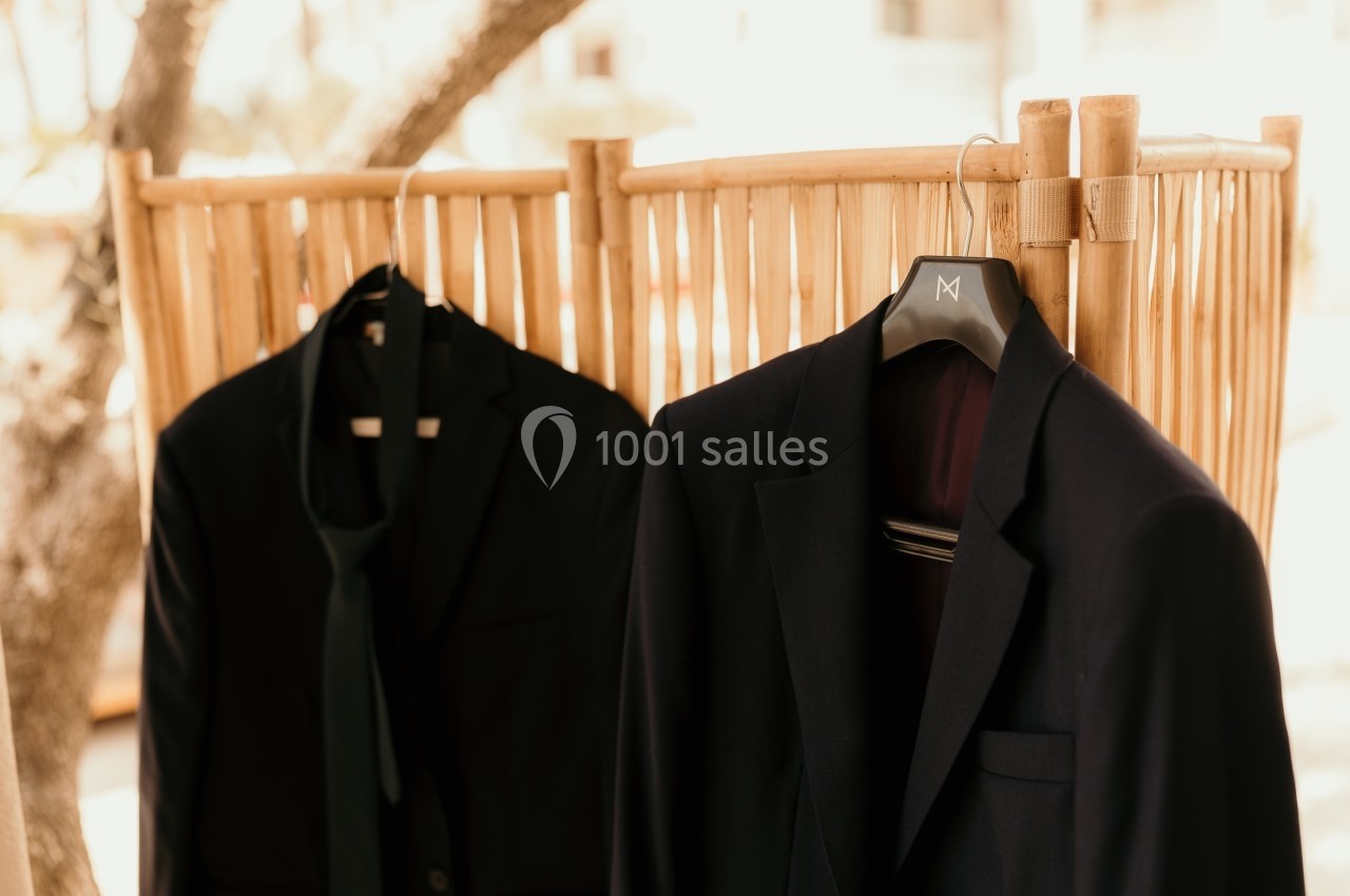 Deux costumes noirs suspendus sur des cintres devant un paravent en bois clair.