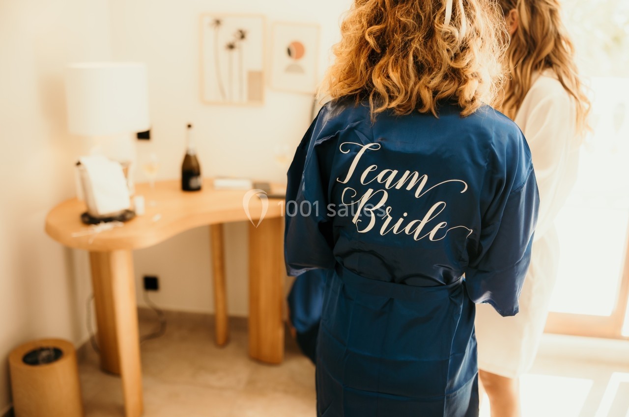 Deux femmes en peignoir, l'une portant un peignoir bleu avec l'inscription ’Team Bride’, dans une pièce lumineuse.
