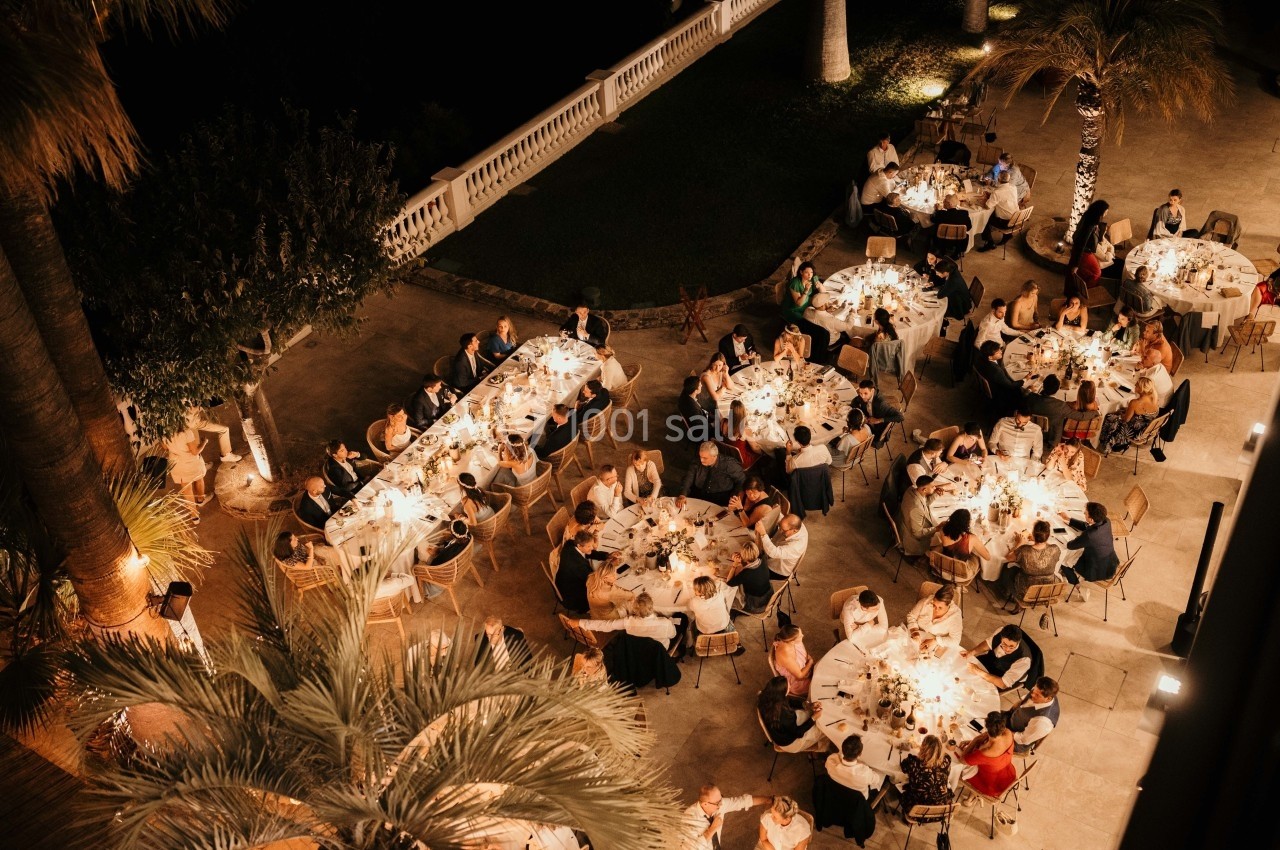 Vue aérienne d'un dîner en plein air avec plusieurs tables rondes et rectangulaires éclairées par des lumières chaleureuses.