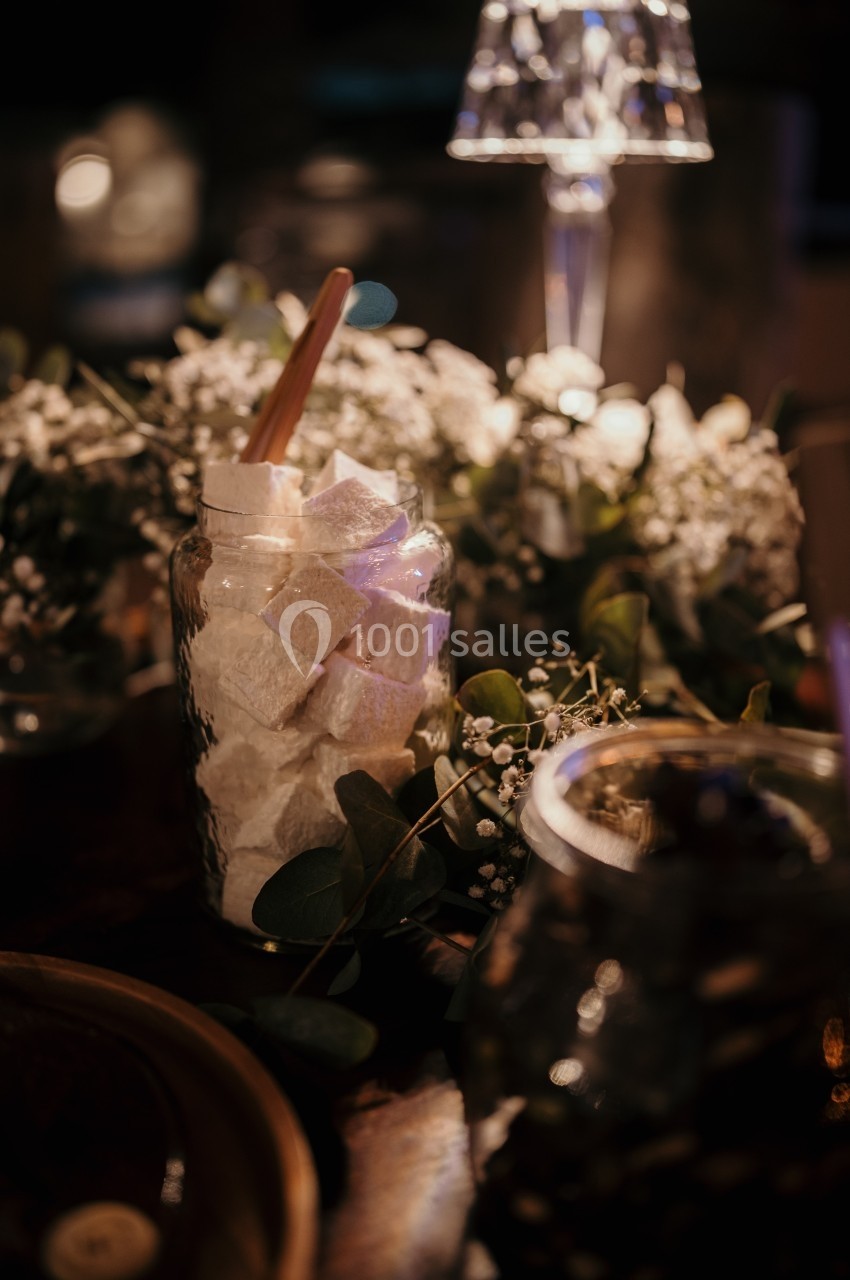 Pot en verre rempli de guimauves blanches, entouré de fleurs et éclairé par une lumière tamisée.