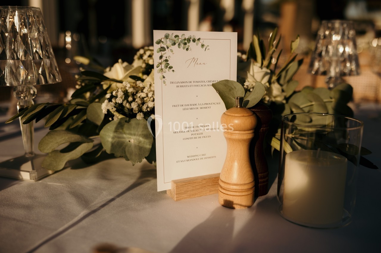Menu de mariage posé sur une table décorée de fleurs, avec un moulin à poivre et une bougie en verre.