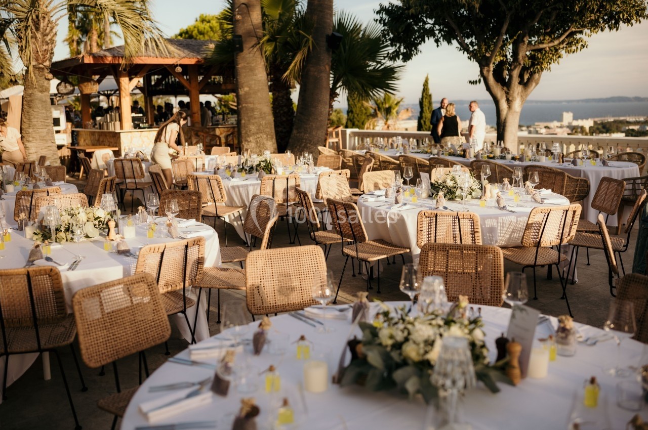 Tables rondes décorées pour un événement en plein air, entourées de chaises en rotin, sous des palmiers.