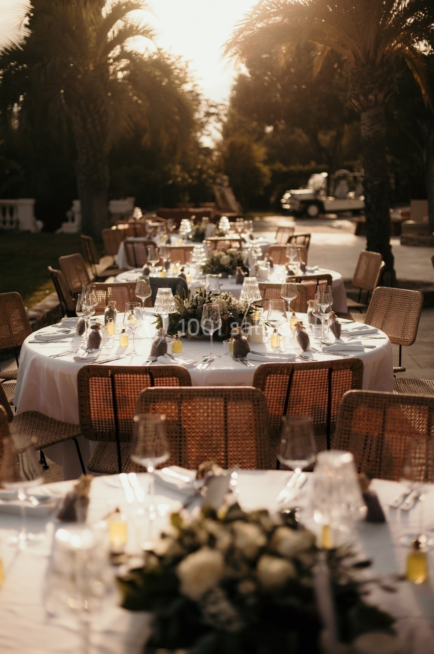Tables élégamment dressées en extérieur avec nappes blanches, verres, bougies et décorations florales, sous des palmiers.