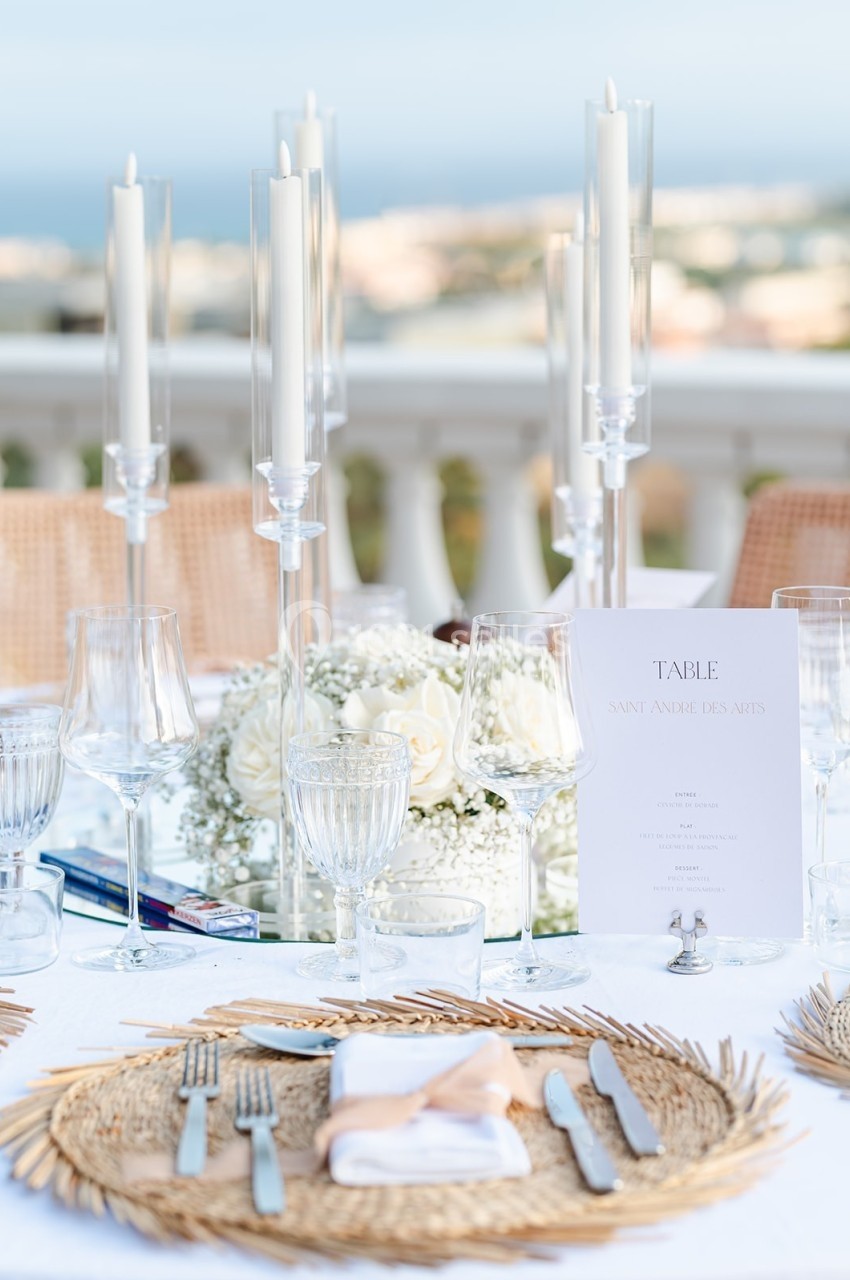Table élégamment dressée en extérieur avec chandeliers, vaisselle raffinée et décoration florale blanche.