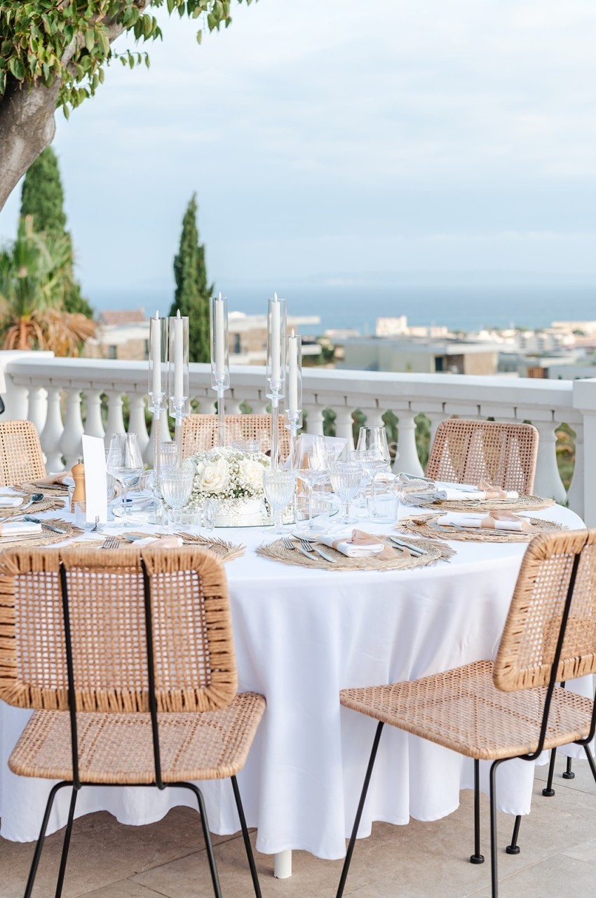 Table dressée en extérieur avec nappes blanches, chaises en rotin, bougies et vue sur un paysage côtier.