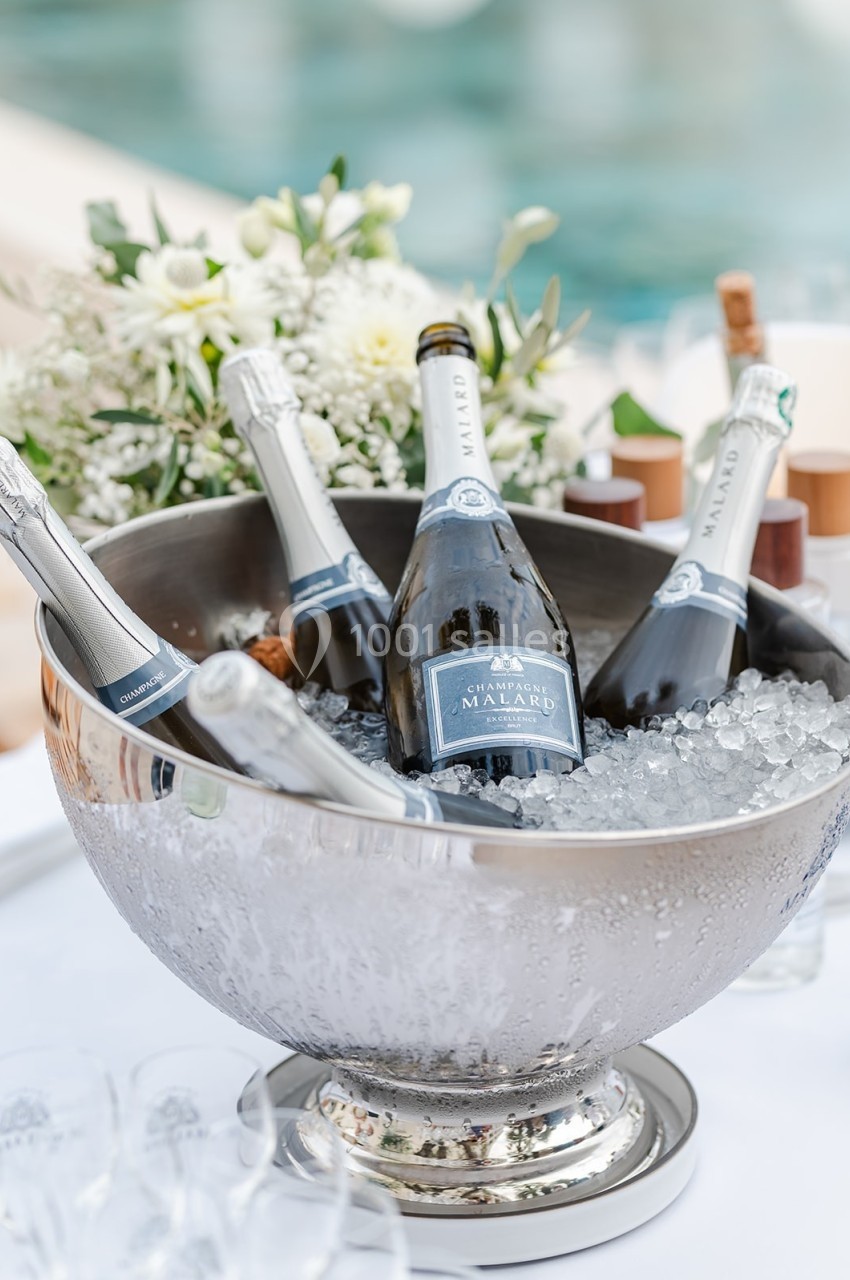 Seau en métal rempli de glace contenant des bouteilles de champagne, avec des fleurs blanches en arrière-plan.