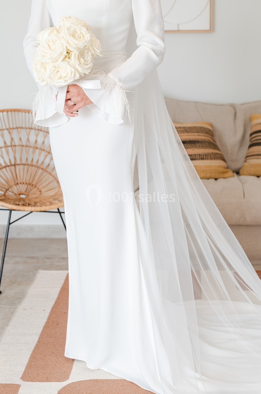 Robe de mariée blanche avec un voile long, tenue par une personne portant un bouquet de roses blanches.