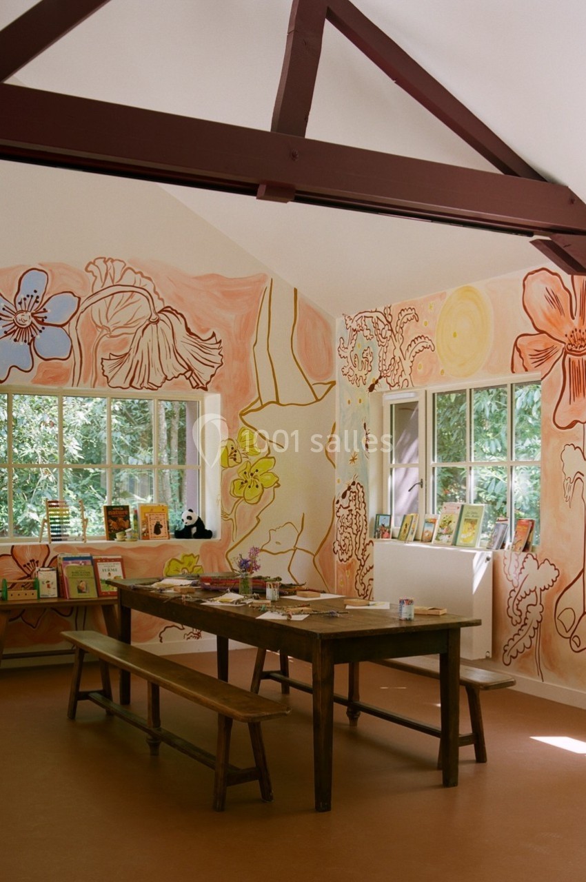 Salle lumineuse avec des murs décorés de peintures florales, tables en bois et fenêtres donnant sur un espace verdoyant.