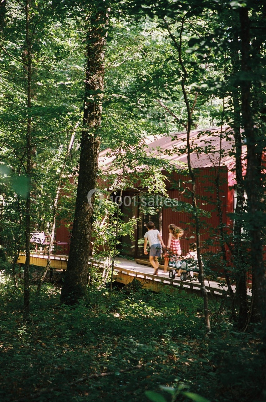 Deux enfants marchent sur une passerelle en bois devant une cabane rouge entourée d'une forêt dense.
