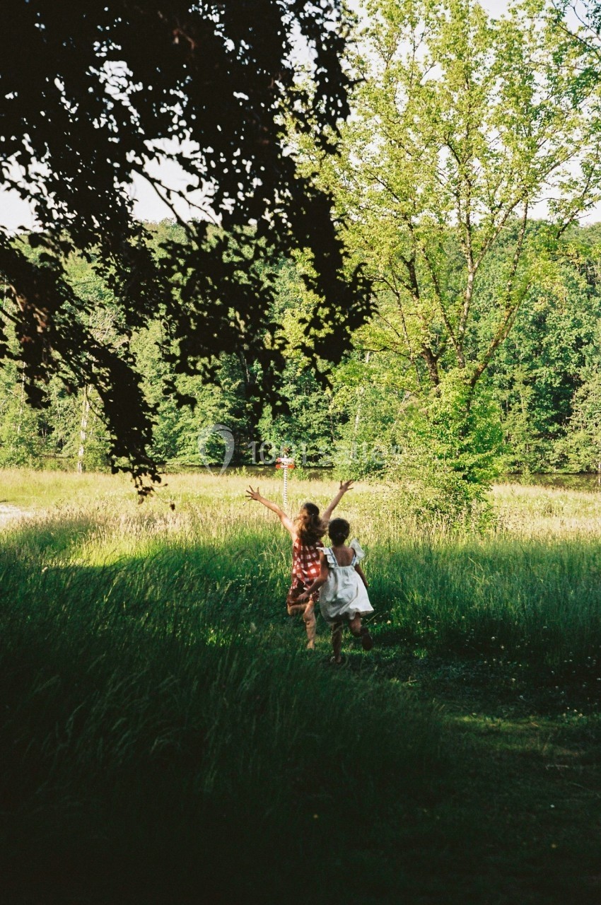 Deux enfants courent dans une prairie verdoyante, entourés d'arbres et baignés par une lumière naturelle.