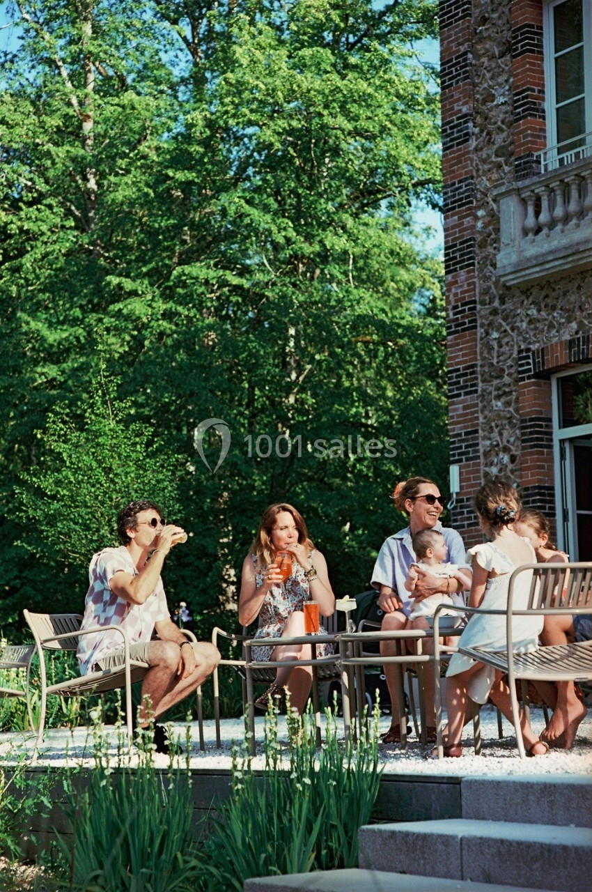 Un groupe de personnes assises autour d'une table en extérieur, près d'un bâtiment en pierre, entouré de verdure.