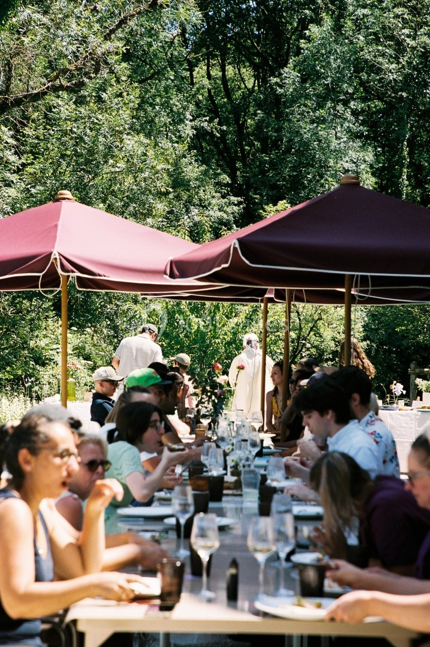 Des personnes déjeunent à de longues tables sous des parasols rouges dans un cadre verdoyant.
