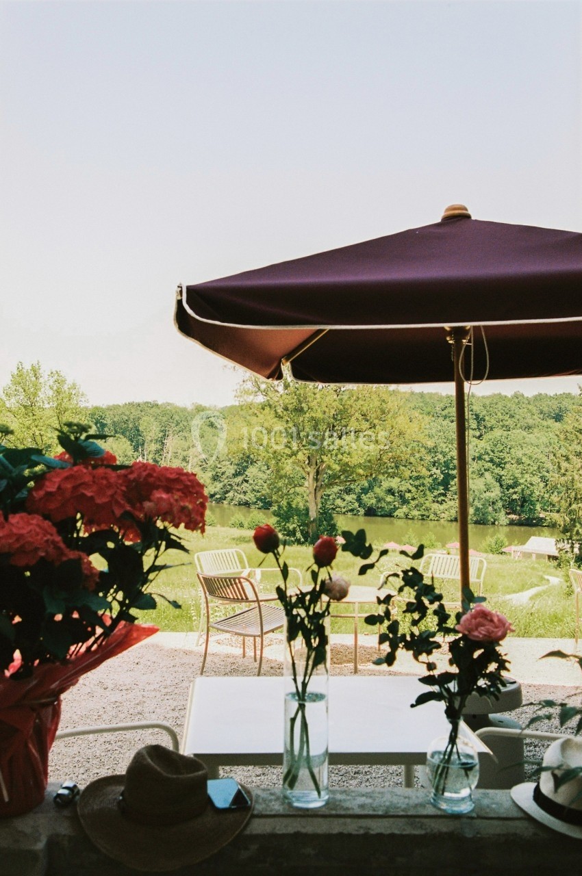 Vue sur un jardin avec des chaises, une table, un parasol et des fleurs en premier plan, près d'un étang.