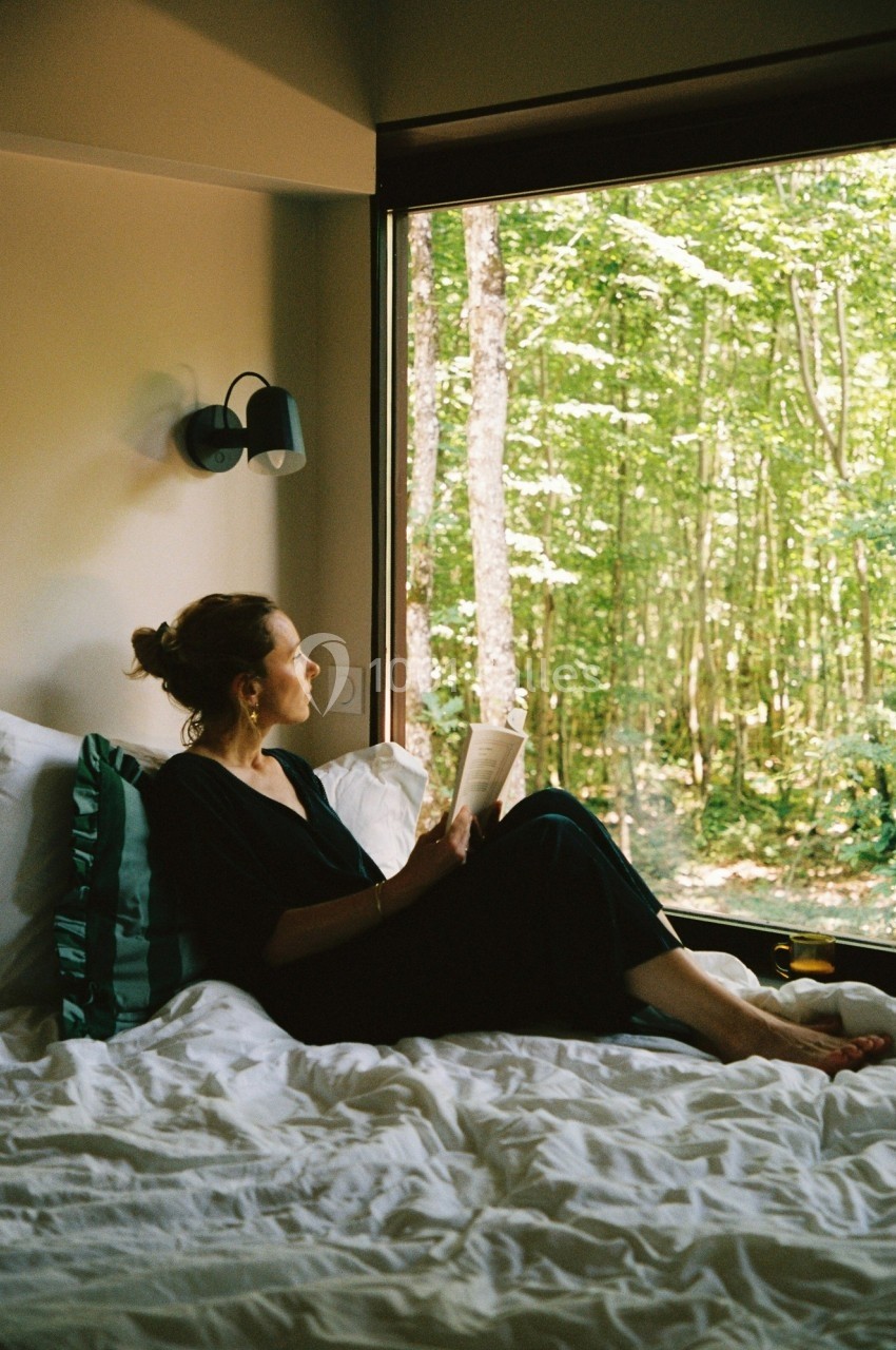 Femme assise sur un lit, lisant un livre près d'une grande fenêtre donnant sur une forêt verdoyante.